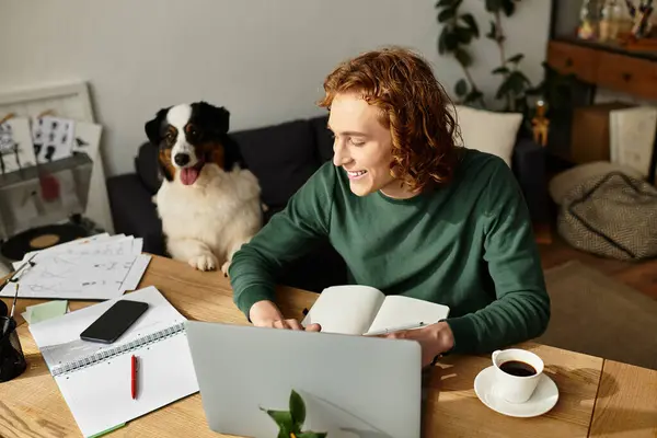 Um jovem senta-se em sua mesa, envolvido no trabalho, enquanto um cão feliz assiste amorosamente nas proximidades. — Fotografia de Stock