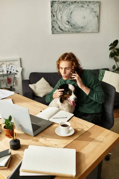 A young man sits at a wooden desk, talking on the phone while his dog snuggles beside him. — Stock Photo