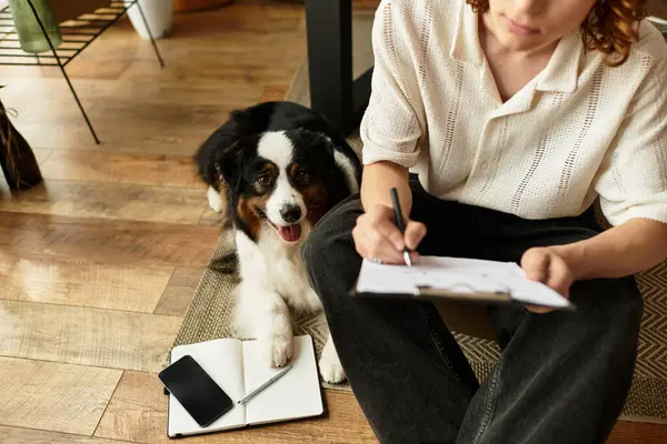 A young man writes in his notebook at home, while his playful dog nearby creates warmth. — Stock Photo