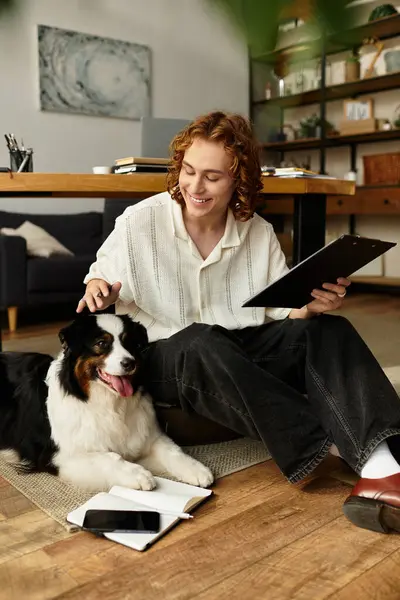 A cheerful young man relaxes at home, taking notes with his dog by his side. — Stock Photo