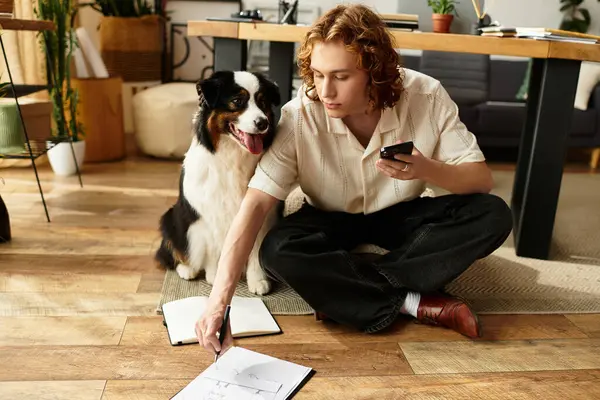 Un joven se sienta cómodamente en el suelo con su perro, participando en material de estudio en casa. - foto de stock
