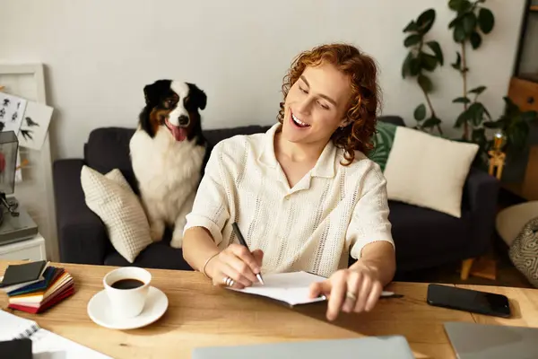 A young man sits at a wooden table in a cozy room, writing notes with his dog nearby. — Stock Photo