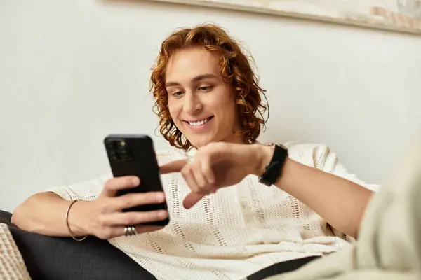 Relajándose cómodamente en casa, un joven se involucra con su teléfono, sonriendo y disfrutando del momento. - foto de stock