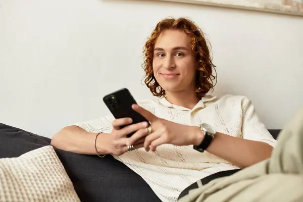 A young man sits on a couch, happily engaged with his smartphone. — Stock Photo
