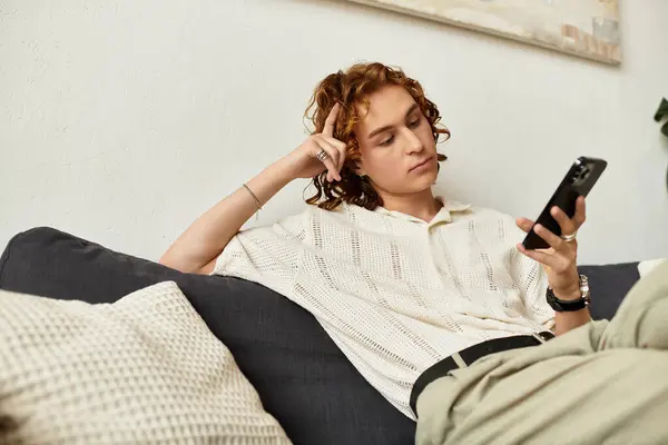 A curly haired young man relaxes on a sofa, engrossed in his smartphone. — Stock Photo
