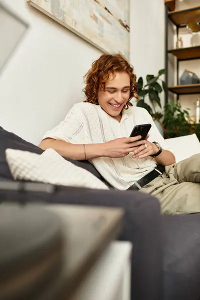 Jovem relaxado senta-se em um sofá, sorrindo enquanto ele interage com seu smartphone em um ambiente acolhedor. — Fotografia de Stock