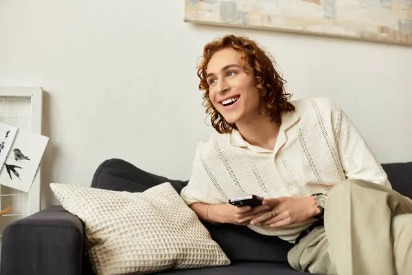 Jovem relaxado com o cabelo encaracolado sorri enquanto assiste TV em um ambiente acolhedor sala de estar. — Fotografia de Stock
