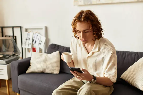 Relaxed young man sits comfortably on a couch, sipping coffee and checking his phone at home. — Stock Photo
