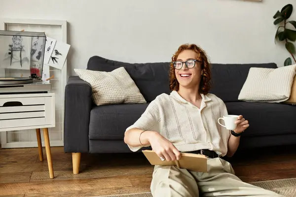 Young man sits comfortably in his cozy living room, holding a warm drink and smiling. — Stock Photo
