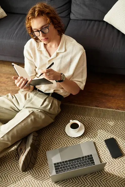Relajado joven se sienta en el suelo en casa, escribiendo en un cuaderno con café cerca. - foto de stock