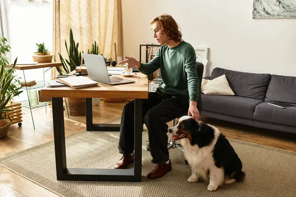 A young man works on his laptop at a desk while his dog relaxes beside him. — Stock Photo
