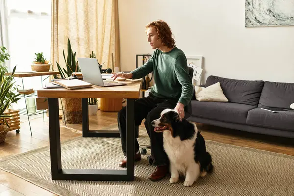 En un acogedor entorno hogareño, un joven se dedica a trabajar en su escritorio acompañado de su alegre perro. - foto de stock