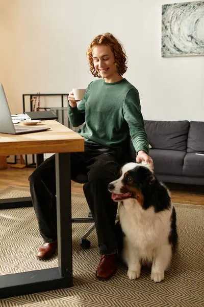 Um jovem senta-se em sua mesa em uma acolhedora sala de estar, tomando café enquanto acaricia seu cão. — Fotografia de Stock
