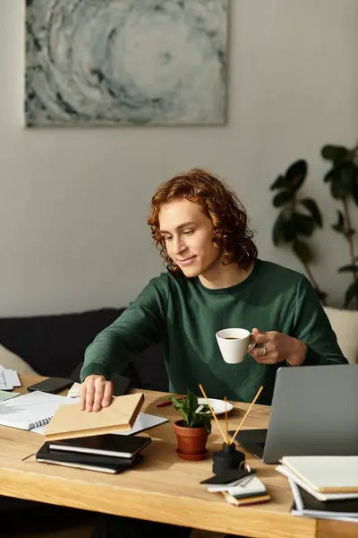 A young man sits comfortably at home, sipping coffee and organizing his workspace with focus. — Stock Photo