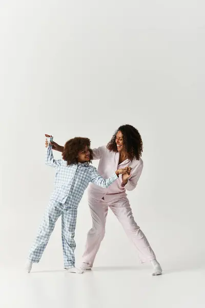 Black mom anddaughter in matching PJs dance joyfully on grey. — Stock Photo