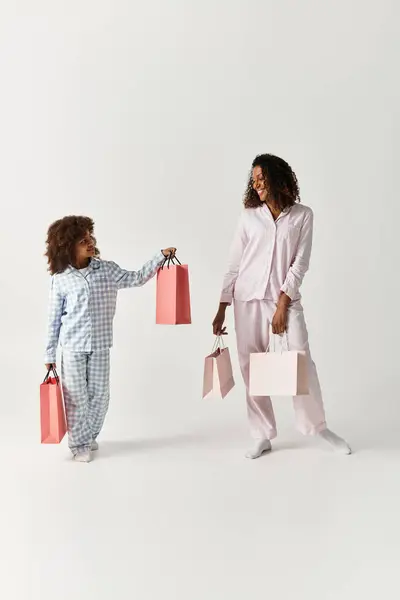 Black mother and daughter in PJs, holding pink shopping bags, grey backdrop. — Stock Photo