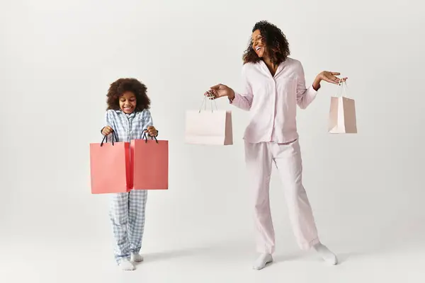 Black mother and daughter in matching PJs smile with shopping bags. — Stock Photo
