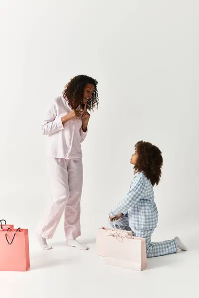 An African American Mother and daughter, both in matching pajamas, pose in a studio next to shopping bags — Stock Photo