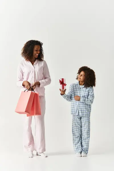 Madre afroamericana che tiene borse della spesa sorride a sua figlia, che le presenta un regalo. — Foto stock