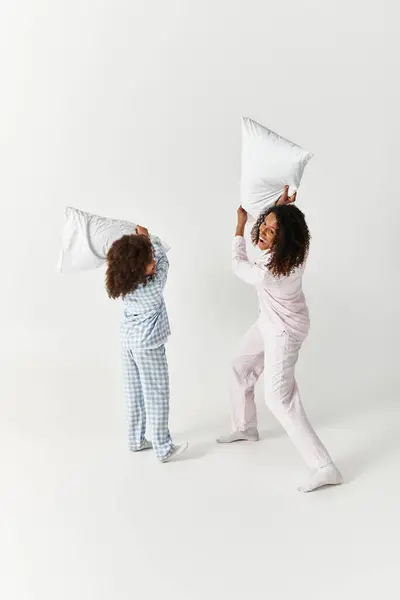 Black mother and daughter having playful pillow fight in PJs on grey. — Stock Photo