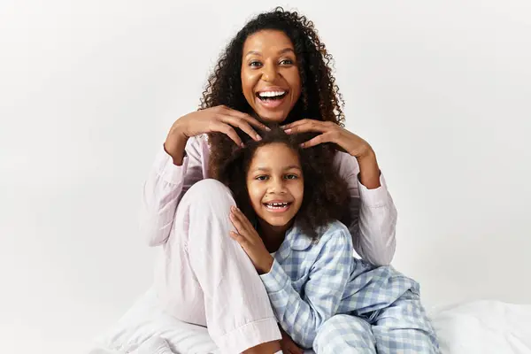 Black mom smiles behind daughter in matching PJs, touching hair. — Stock Photo