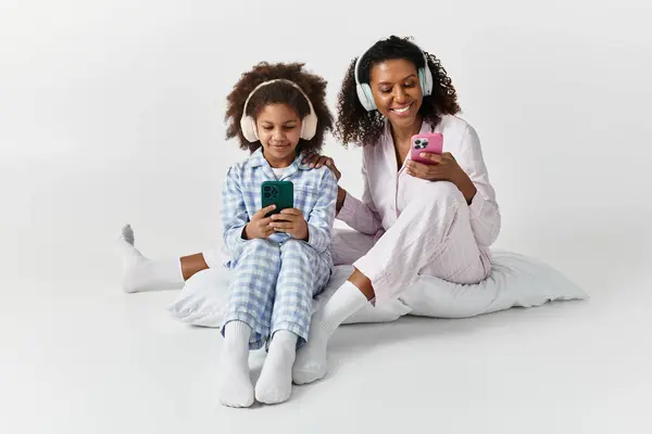 Black mom and daughter in PJs and headphones use phones on grey. — Stock Photo