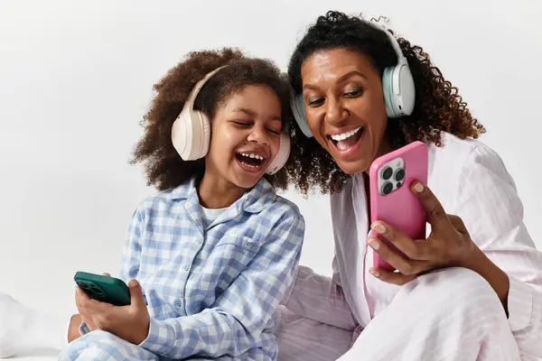 African American mother and daughter are listening to music on their phones in matching pyjamas. — Stock Photo