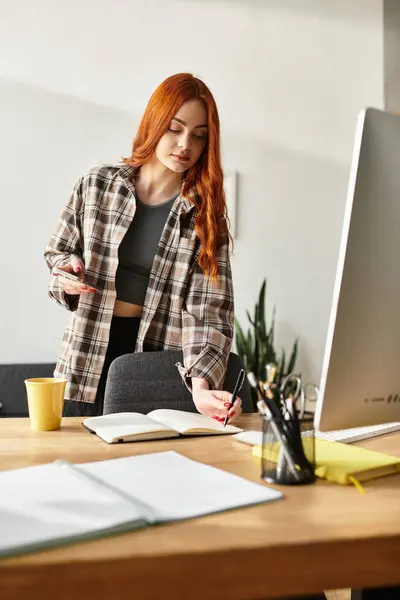 Una joven con el pelo rojo largo escribe en su cuaderno en su acogedora oficina en casa. - foto de stock