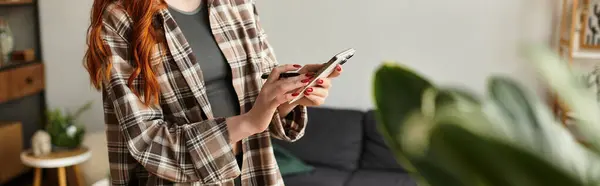 Woman with long red hair relaxes at home while texting on her phone, surrounded by greenery. — Stock Photo