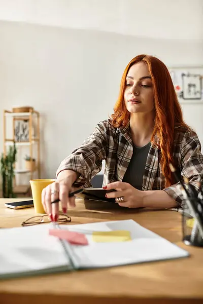 A young woman sits at a wooden table, creating with notes and a drink close by. — Stock Photo