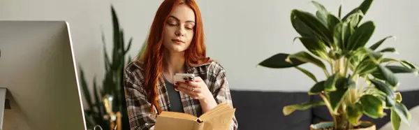 A young woman with red hair reads a book while checking her phone in a stylish interior. — стоковое фото