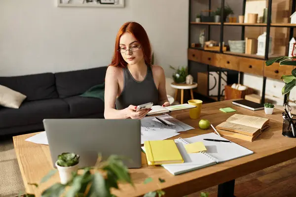 Woman with red hair and glasses focuses on her phone while studying in a cozy workspace. — Stock Photo