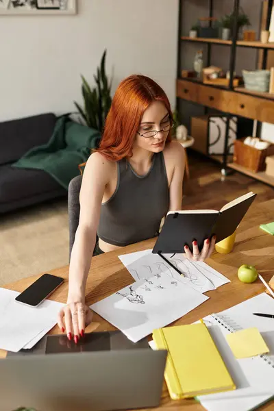Beautiful woman with red hair focused on reading a book while surrounded by papers and a laptop — Stock Photo