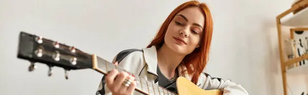 Молодая женщина с рыжими волосами улыбается, играя на гитаре в теплом и уютном месте. — стоковое фото