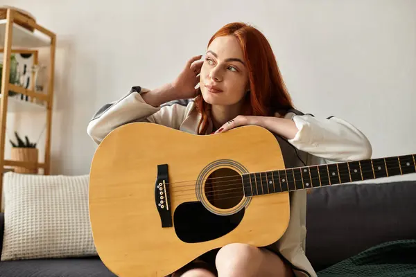A young beautiful woman plays her guitar while relaxing on a comfortable sofa in a serene room. — Stock Photo