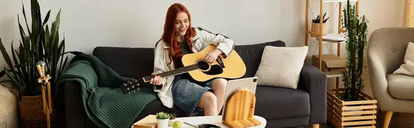 Giovane donna con i capelli rossi strums una chitarra mentre seduto comodamente nel suo spazio di vita. — Foto stock