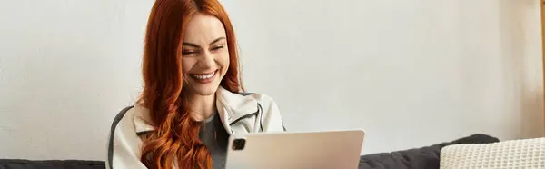 A cheerful young woman with red hair smiles while exploring content on her tablet at home. — Stock Photo