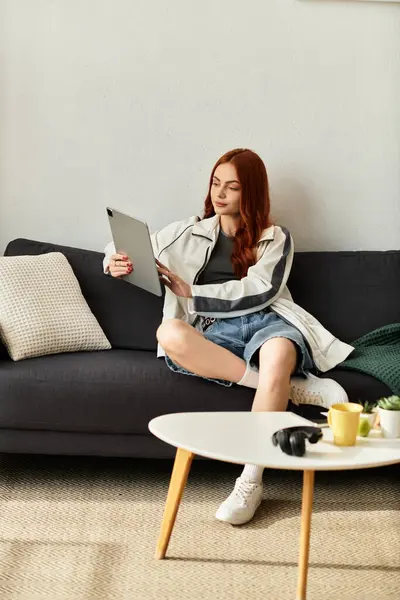 Jeune femme aux longs cheveux roux s'assoit confortablement sur le canapé, absorbé dans sa tablette. — Photo de stock