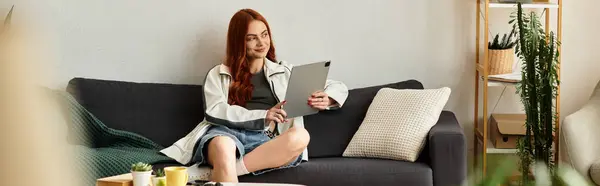 Una giovane donna dai lunghi capelli rossi si rilassa su un divano, impegnandosi con il suo tablet in uno spazio accogliente. — Foto stock