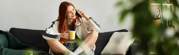 Молодая женщина сидит на диване, с радостью потягивая кофе и разговаривая по телефону. — стоковое фото