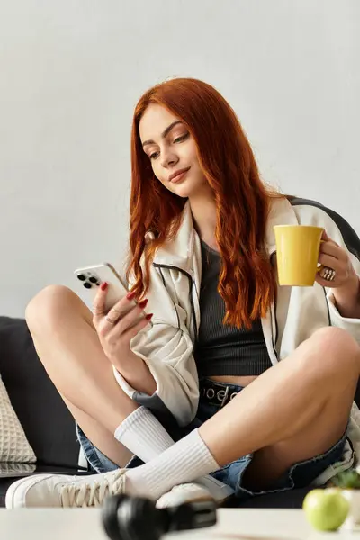 A young woman relaxes on a couch, sipping from a yellow mug and exploring her phone. — Stock Photo