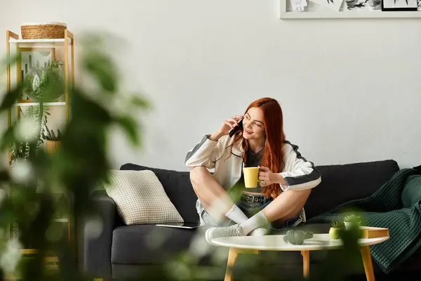 Giovane donna comodamente si siede su un divano, chiacchierando al telefono mentre si gode una bevanda calda. — Foto stock
