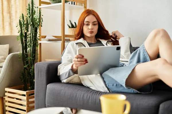 Una giovane donna attraente si rilassa su un divano, concentrata sul suo tablet sorseggiando una bevanda calda. — Foto stock