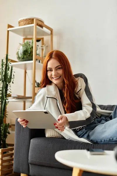 Smiling young woman with red hair comfortably lounges on a sofa, engaging with her tablet. — Stock Photo