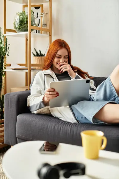 Una giovane donna con bei capelli rossi sorride mentre è impegnata con il suo iPad in un ambiente confortevole. — Foto stock