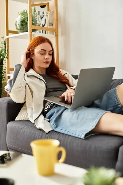 A young beautiful woman calmly types on her laptop while lounging comfortably on a sofa. — Stock Photo