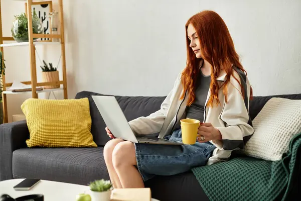 A young woman with red hair relaxes on a couch, sipping from a yellow mug and using a laptop. — Stock Photo