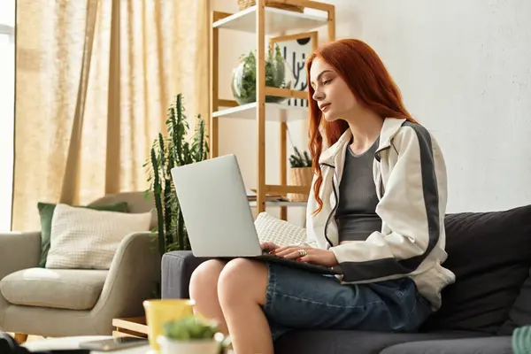 A young woman with red hair focused on her laptop in a cozy living space, relaxing indoors. — Stock Photo