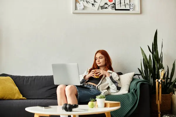 A young beautiful woman relaxes on a sofa, sipping tea and browsing her laptop happily. — Stock Photo