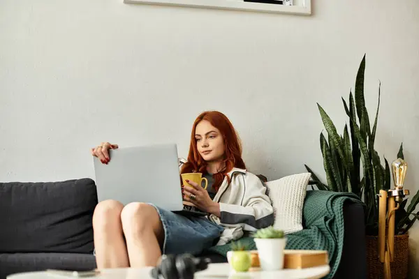 Relaxed young woman sits on a couch, sipping a warm drink and using her laptop in a cozy space. — стоковое фото